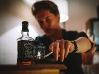 A moody image of a man smoking with a glass of whiskey indoors.