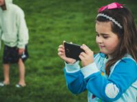 A young girl with a smartphone taking a photo in a grassy park setting