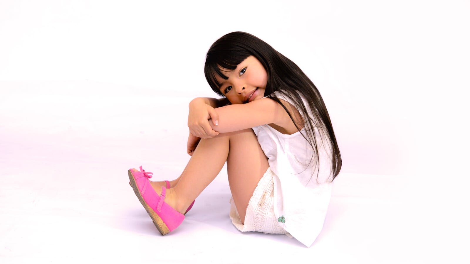A cheerful young girl sitting on a white backdrop wearing bright pink shoes and a summer dress.