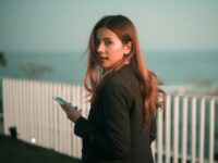 Stylish young woman holds smartphone with a scenic sea backdrop outdoors.