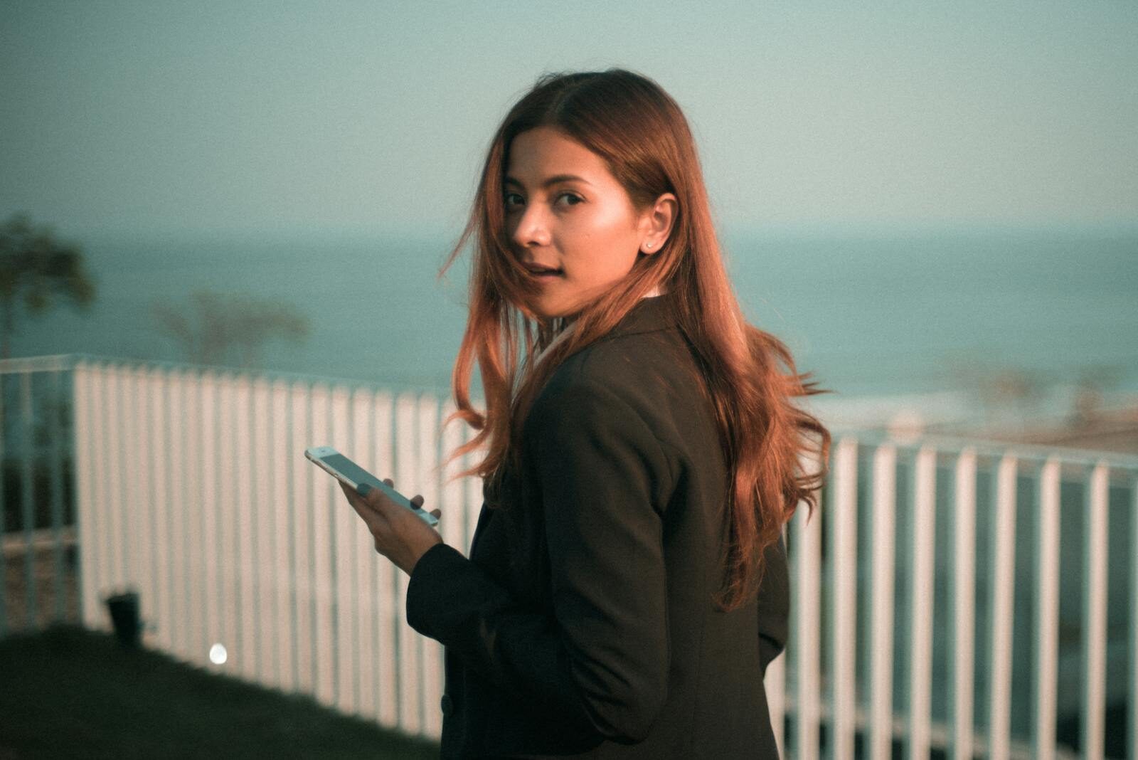 Stylish young woman holds smartphone with a scenic sea backdrop outdoors.
