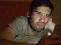 An adult man in a dim room looking at a smartphone with a thoughtful expression.