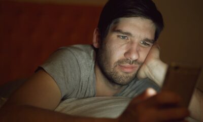 An adult man in a dim room looking at a smartphone with a thoughtful expression.