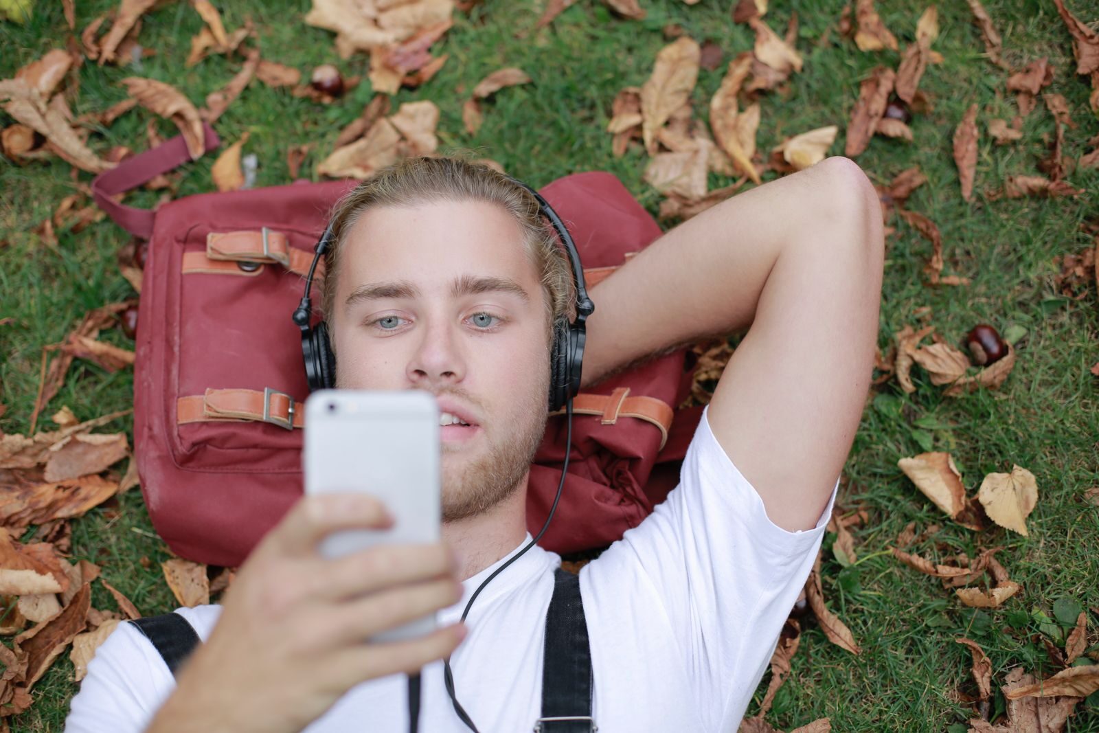 A young man lying on grass with headphones, enjoying music and using his smartphone in a park.