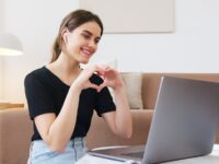 Cheerful happy young female in jeans and black shirt having conversation via video call on laptop and making heart shape with fingers while sitting on floor in modern living room