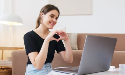 Cheerful happy young female in jeans and black shirt having conversation via video call on laptop and making heart shape with fingers while sitting on floor in modern living room