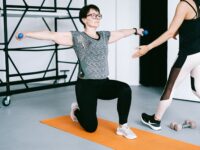 Two women working out with dumbbells on yoga mats in a modern gym setting, focusing on strength training.