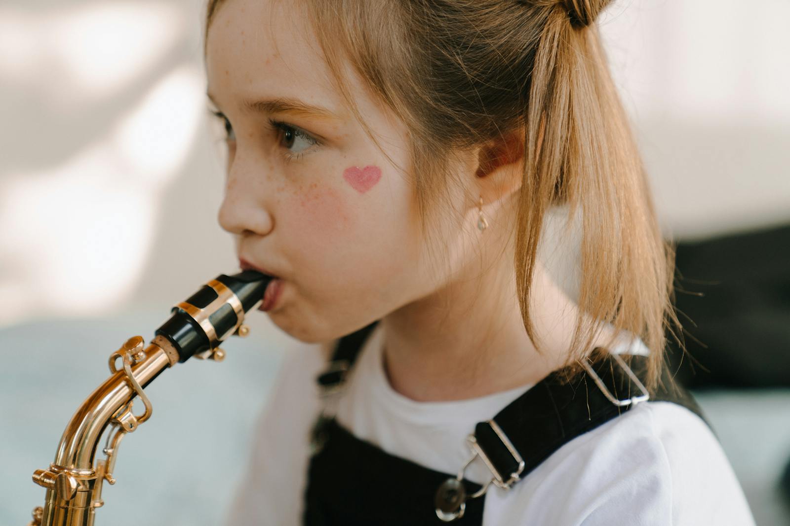 A young girl with heart face paint plays the saxophone indoors, exuding youthful creativity.