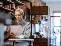 Low angle of concentrated female sitting at table with cup of coffee and reading book at home