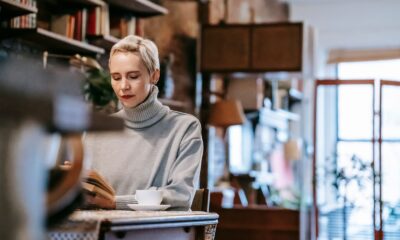 Low angle of concentrated female sitting at table with cup of coffee and reading book at home