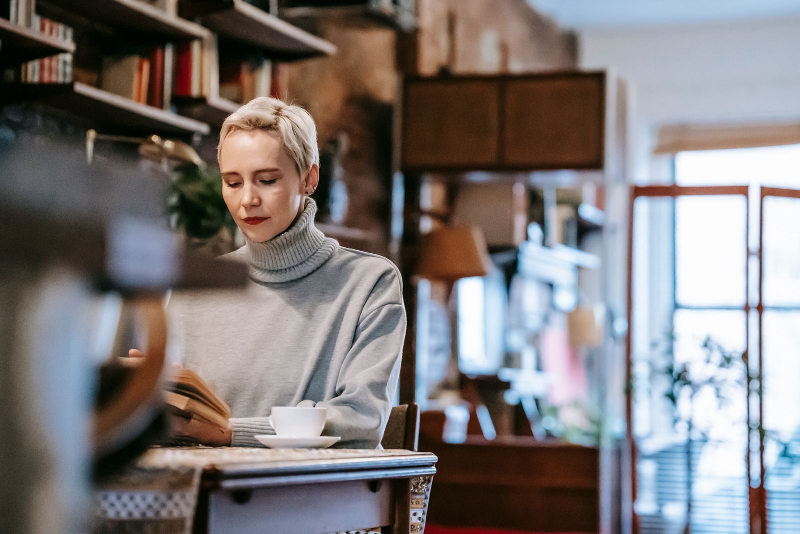 Low angle of concentrated female sitting at table with cup of coffee and reading book at home