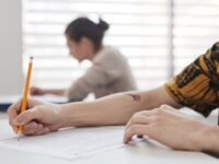 Close-up of student's hands writing on exam sheet, indoors with blurred background.