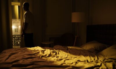 Man in dark bedroom gazing out window, highlighting themes of insomnia and solitude.