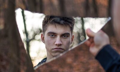 A striking portrait of a man reflected in a broken mirror, set against a forest background.