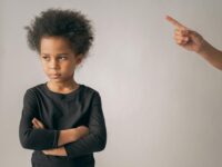 Crop anonymous person pointing up with index finger while scolding unhappy African American girl looking away while standing on white background in studio