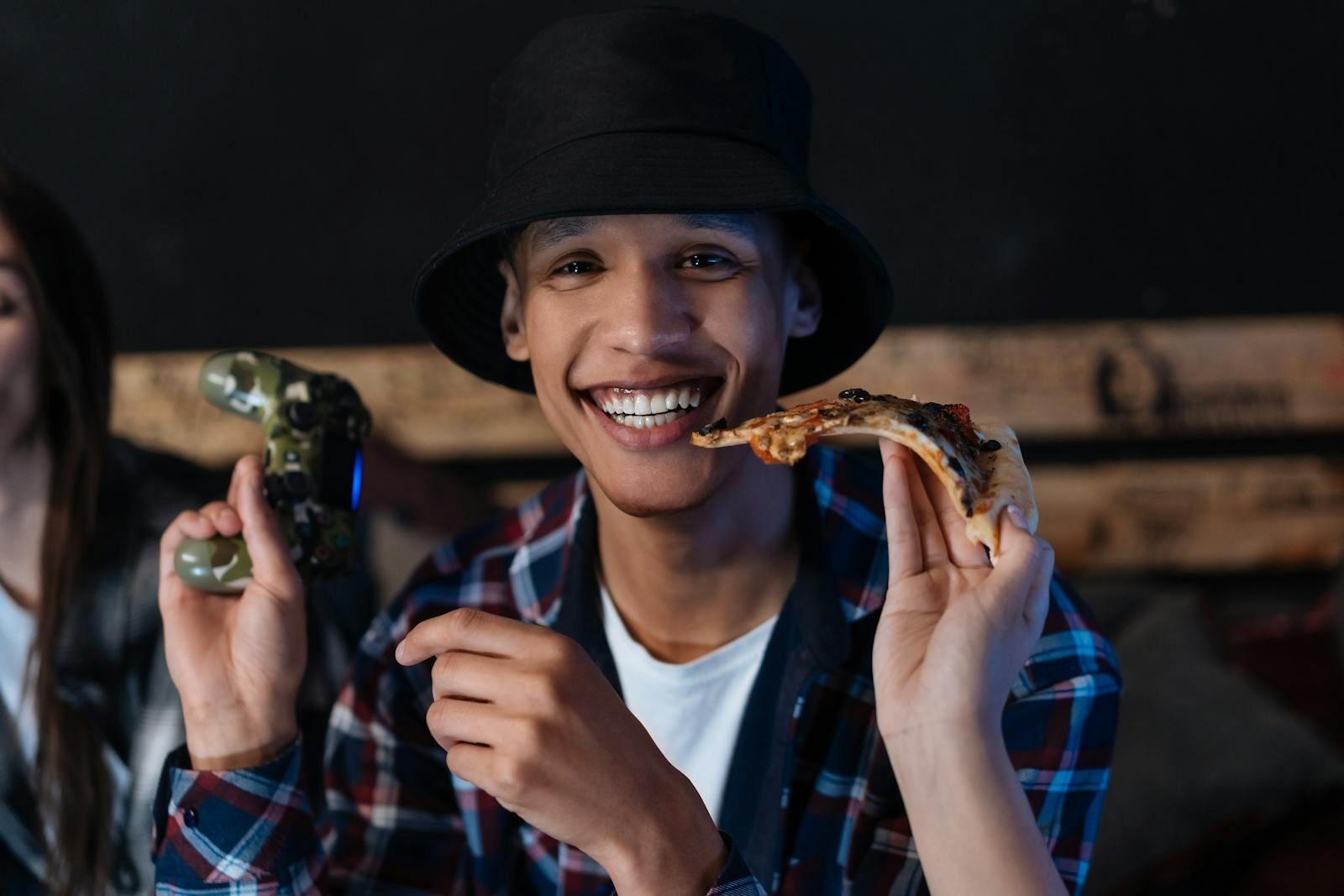 Young man smiling with pizza and joystick indoors, capturing a fun gaming moment.