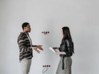 A man and woman in stylish outfits having a discussion in an interior setting with a white wall.