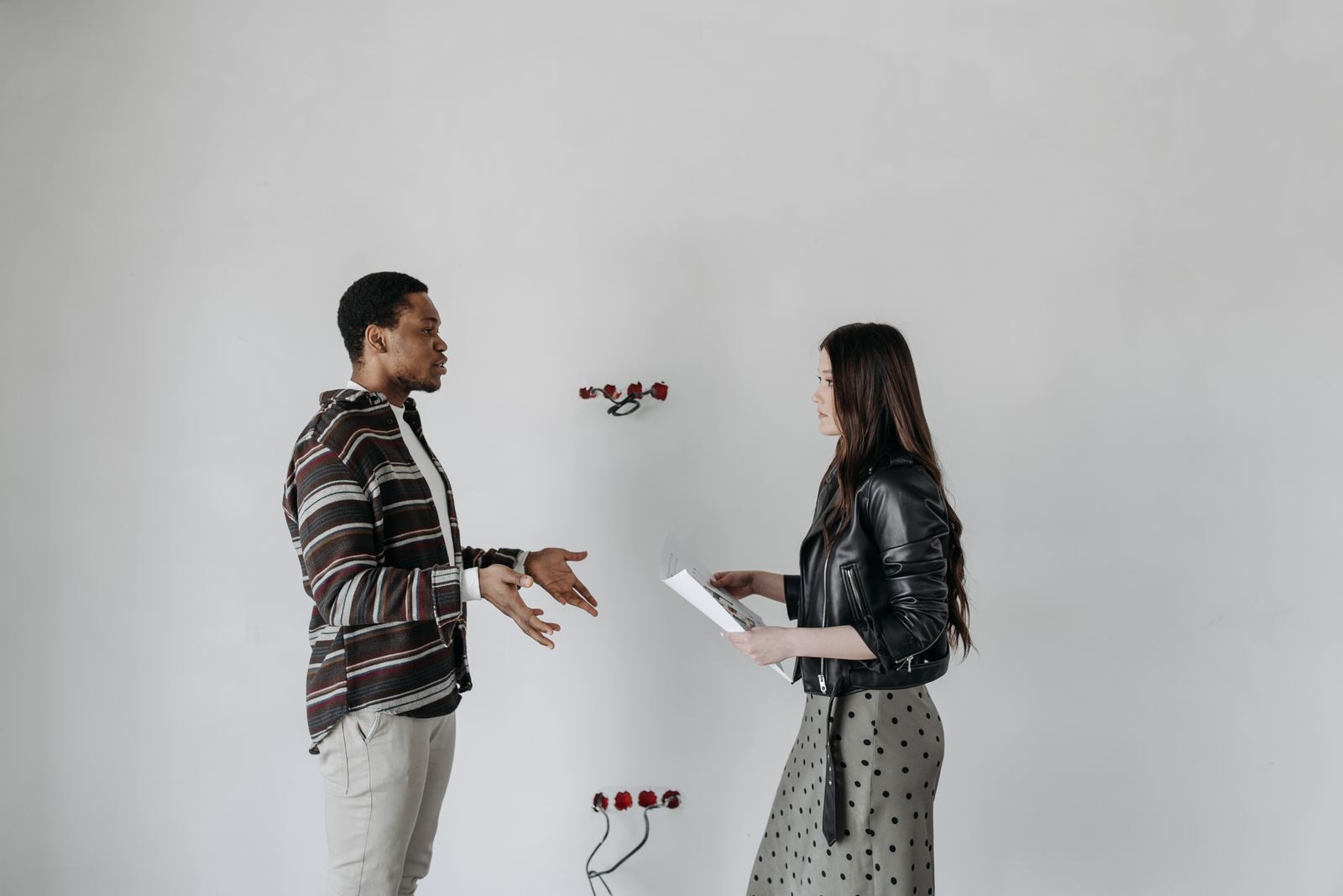 A man and woman in stylish outfits having a discussion in an interior setting with a white wall.