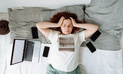 A distressed woman lying in bed surrounded by digital devices, showing signs of stress.