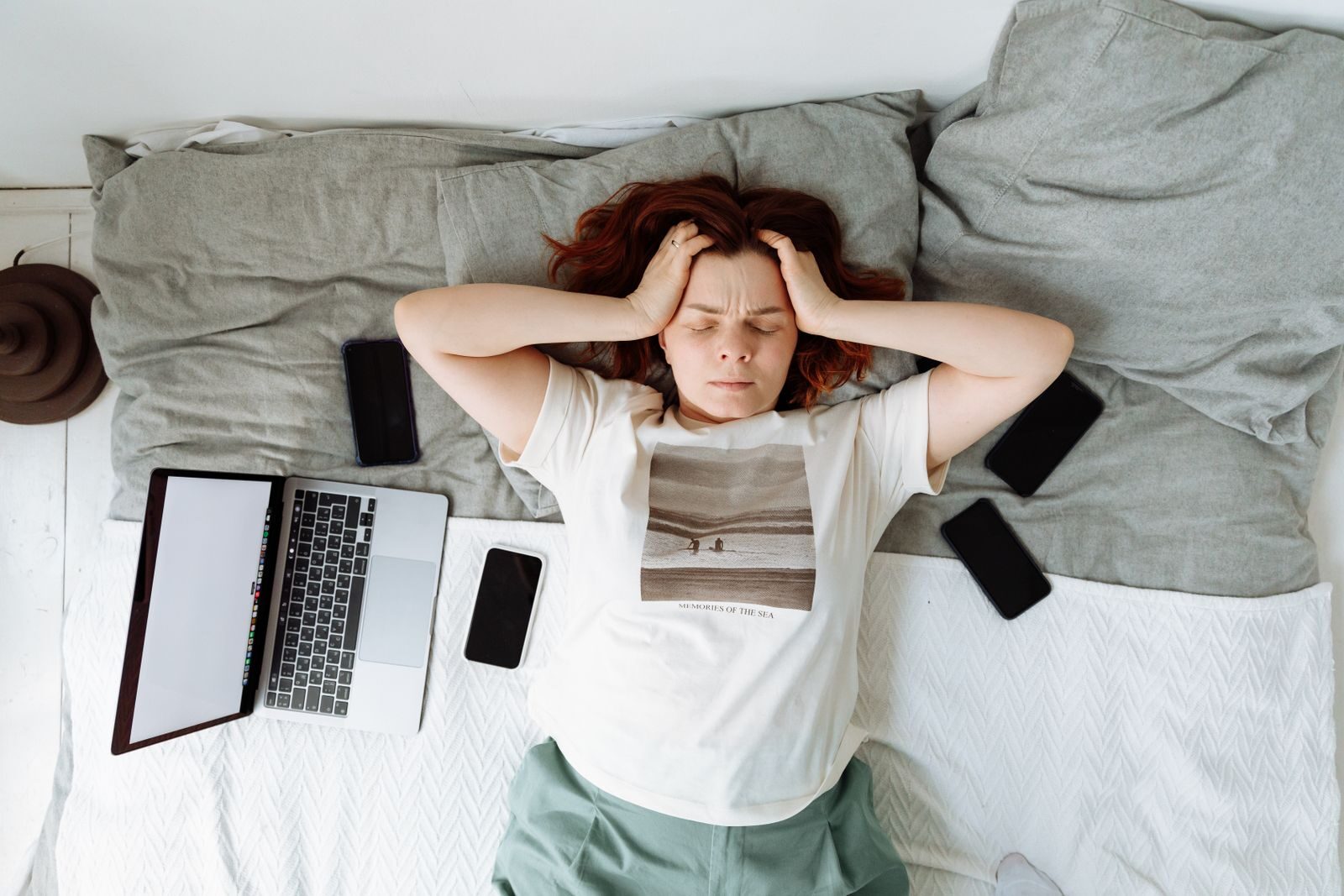 A distressed woman lying in bed surrounded by digital devices, showing signs of stress.