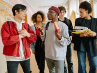 A group of diverse students walking and talking in a school corridor, carrying books and folders.