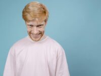 Young man in pink shirt smiles mischievously against blue background in studio setting.