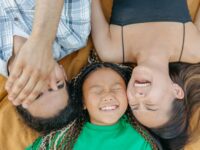 A joyful family bonding moment with parents and daughter laughing while lying on a blanket outdoors.