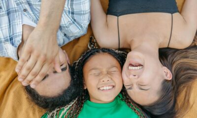 A joyful family bonding moment with parents and daughter laughing while lying on a blanket outdoors.